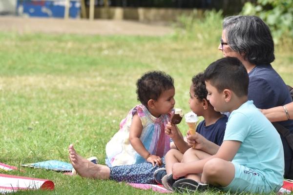 Three small children and an adult eat ice creams while sat on a picnic blanket in Barra Hall Park on a sunny day.
