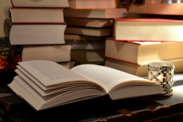Photo of open book on desk with piles of books behind it.