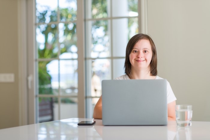 Photo of young woman with Downs Syndrome, sat working at a laptop, smiling