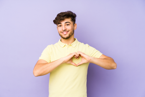 Photo of a young man with his hands together in a shape of a heart, on a light purple background